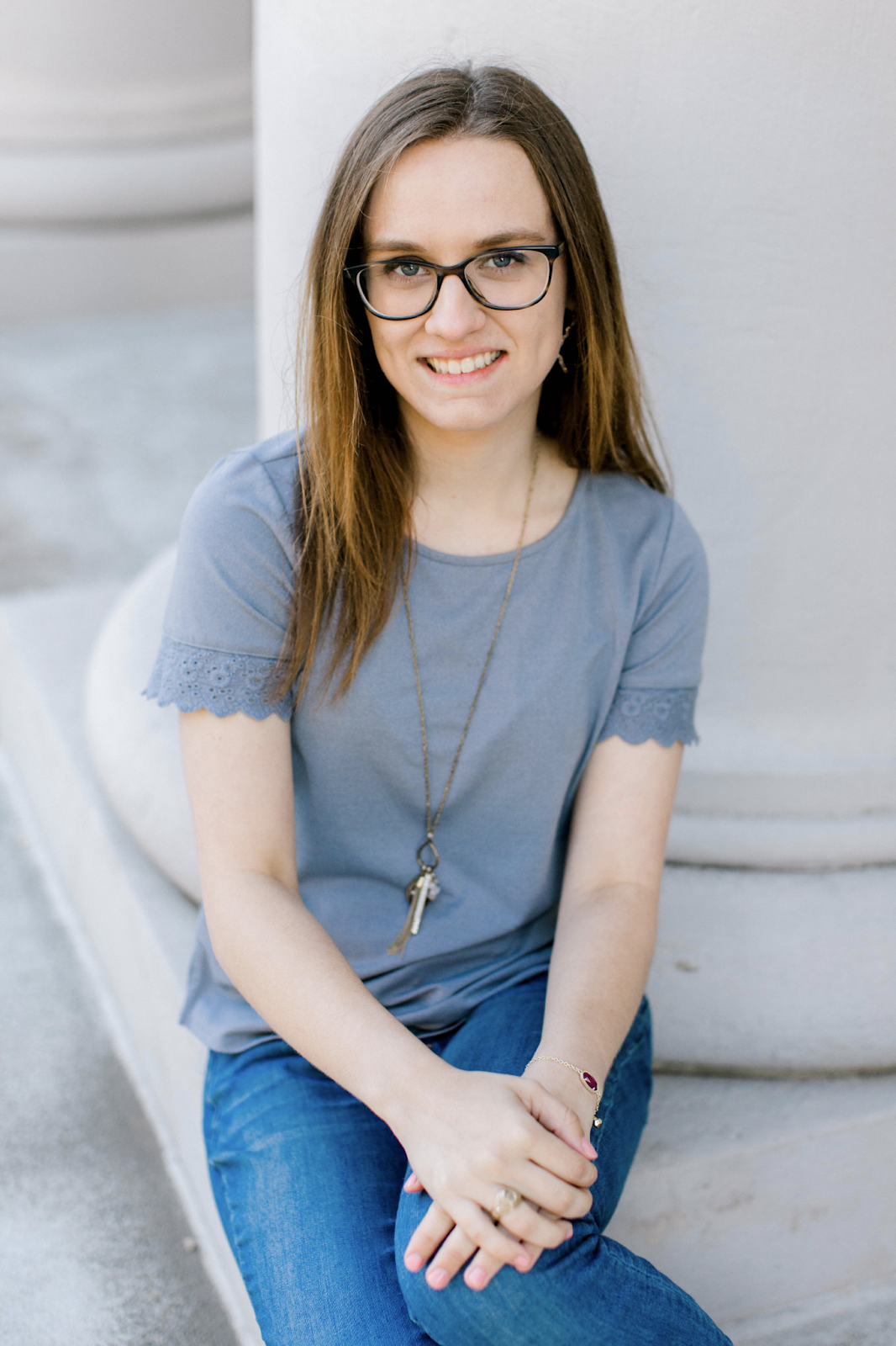 Darby Salge, a white woman in her twenties, is sitting and smiling at the camera. She is wearing brown glasses, a blue top, and blue jeans.