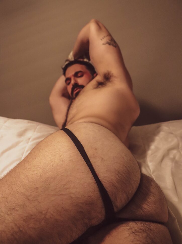 Thick Macho lying down on the bed in a black jockstrap showing off his hairy ass cheeks and smirking down at the camera