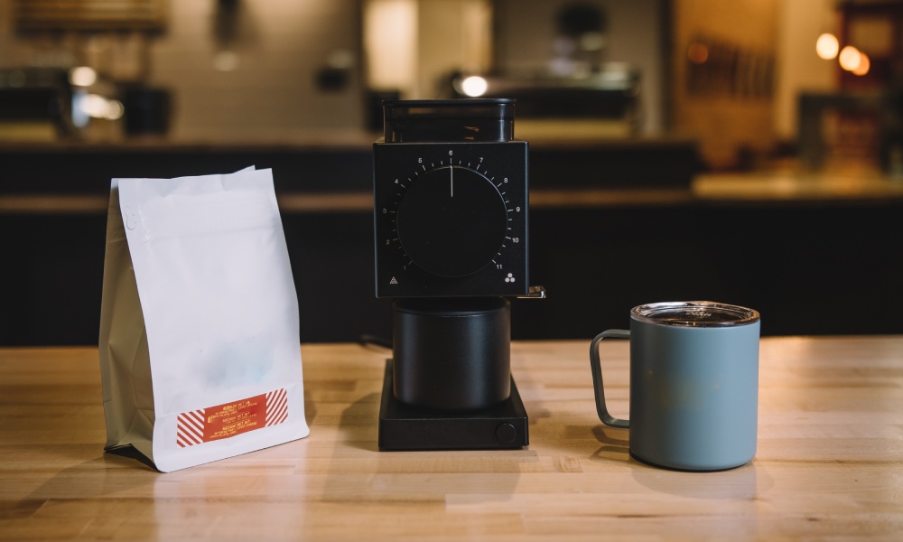 White multilayer kraft paper coffee bag with red and yellow label on wooden counter beside a coffee grinder and blue mug. 