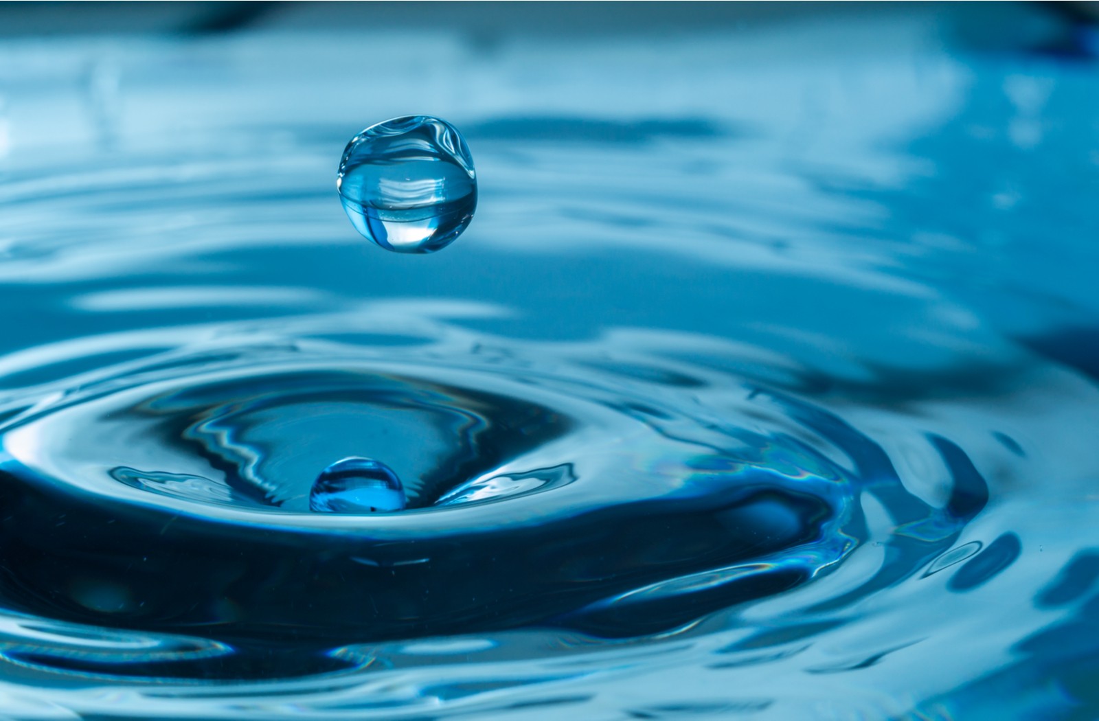 Zoomed perspective of a single blue spherical water drop about to fall into a body of water that is already rippling