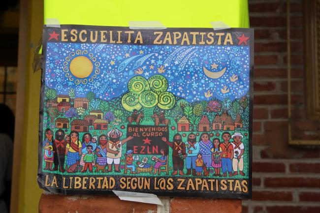 A painted sign for the Escuelita Zapatista, it depicts a diverse group of people pointing to an EZLN sign, with a backdrop of modest houses, a landscape, and the stars.