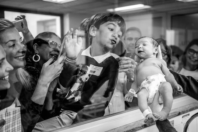 Menino beija o vidro que a sapara da irmã recém nascida. Ela é mostrada a familiares e amigos