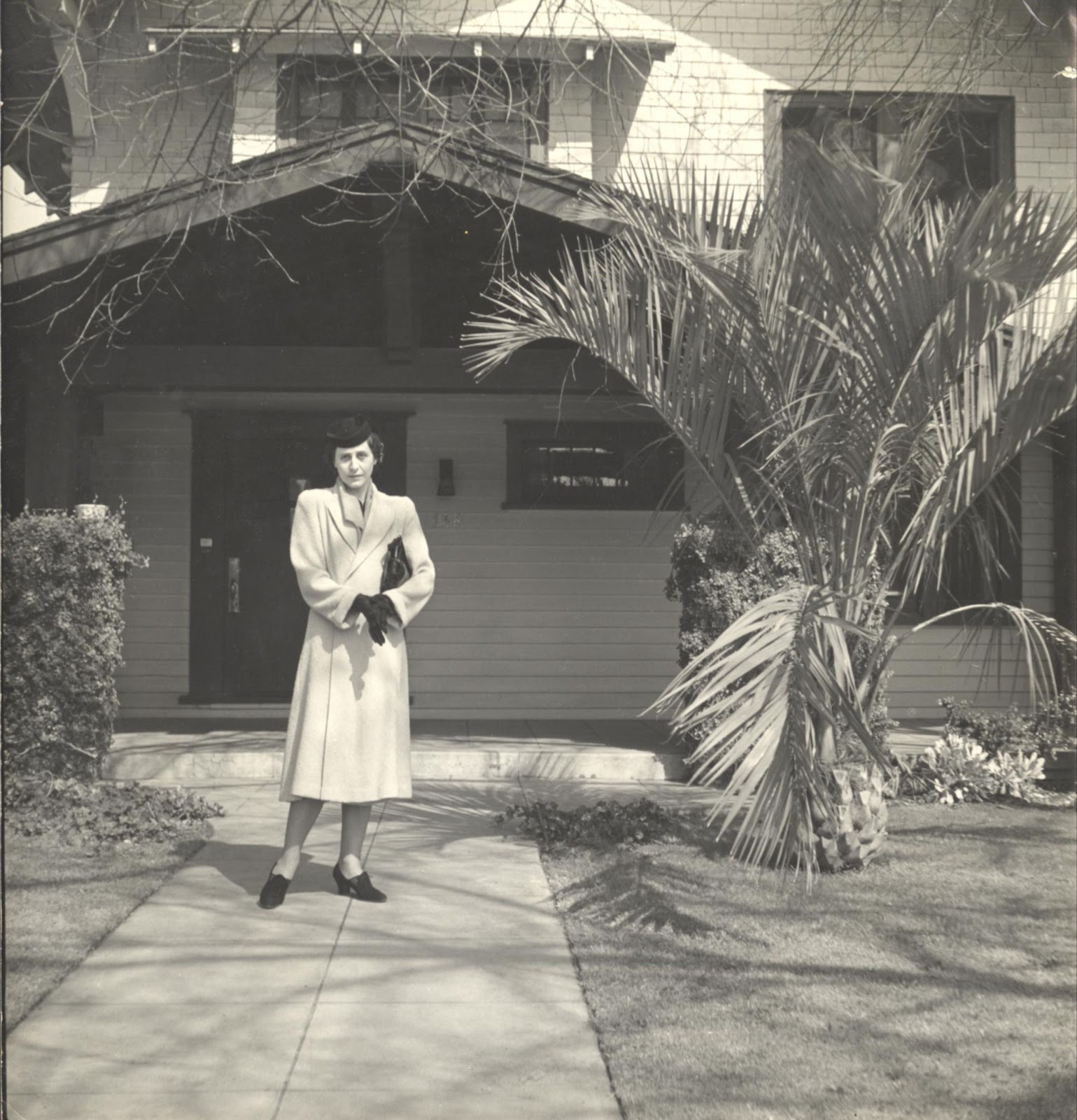 Sepia image of a woman wearing a long coat and dark gloves, tucking a bag under her arm, in front of a house next to a short palm tree.