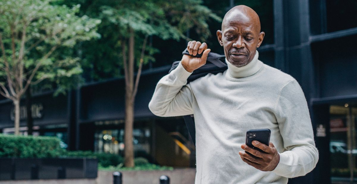 A smiling dark-skinned male customer in a grey sweater carrying a bag over his right shoulder with a smartphone in his left hand as he reads a delightful Ramadan message from his favorite brand.