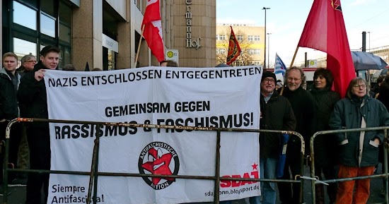 Protestierende mit roten Fahnen und Plakat: »Nazis entschlossen entgegentreten. Gemeinsam gegen Rassismus & Faschismus! Siempre Antifascista! Antifaschistische Aktion. SDAJ«.