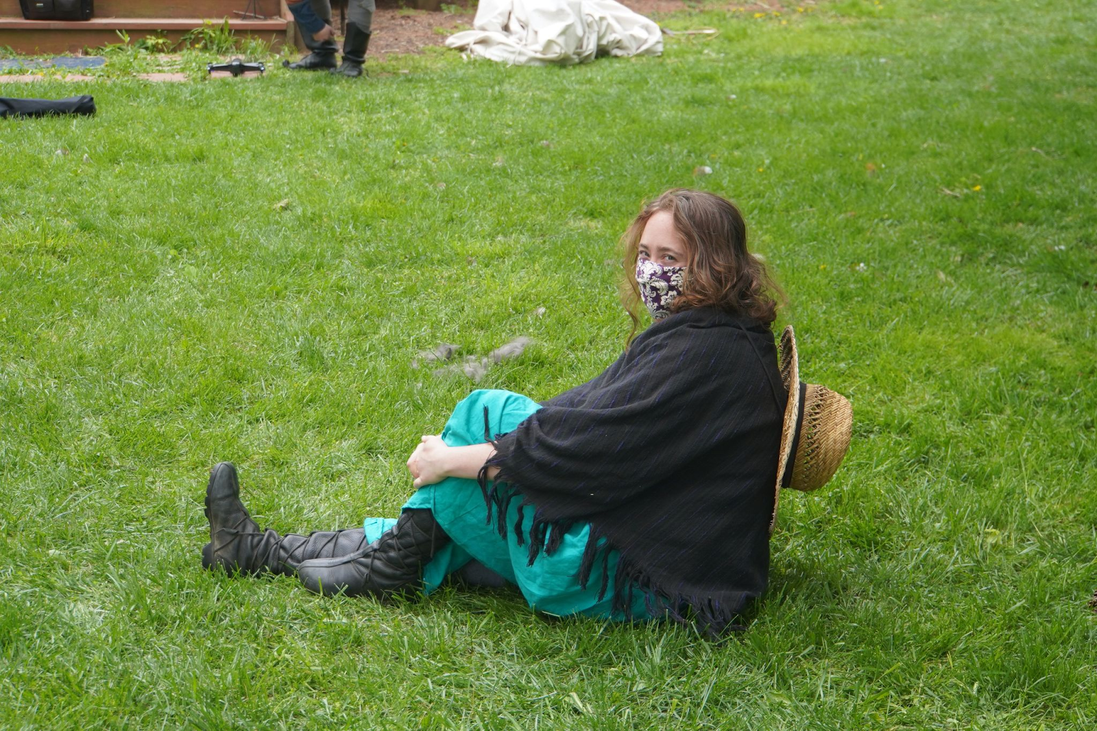 A person sits profile on the grass, one leg stretched out in front of them, but head turned to face the camera. They are wearing black boots, a tea dress, a black shawl, a purple patterned mask, and have a straw hat on their back.