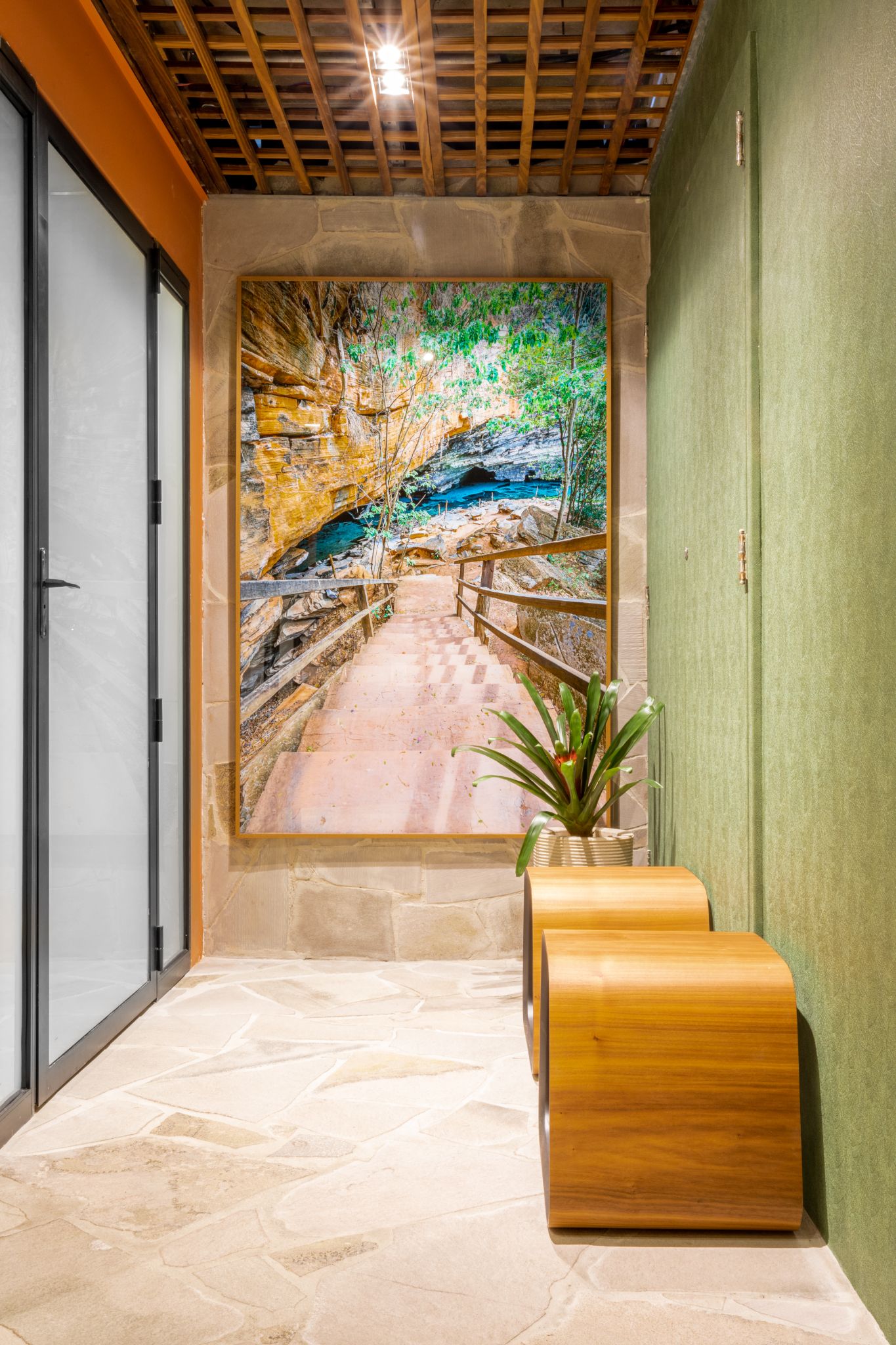 Banheiro com papel de parede imitando folhagens, piso e parede central revestidos de pedra basalto cinza, bancos de madeira e quadro grande decorativo.
