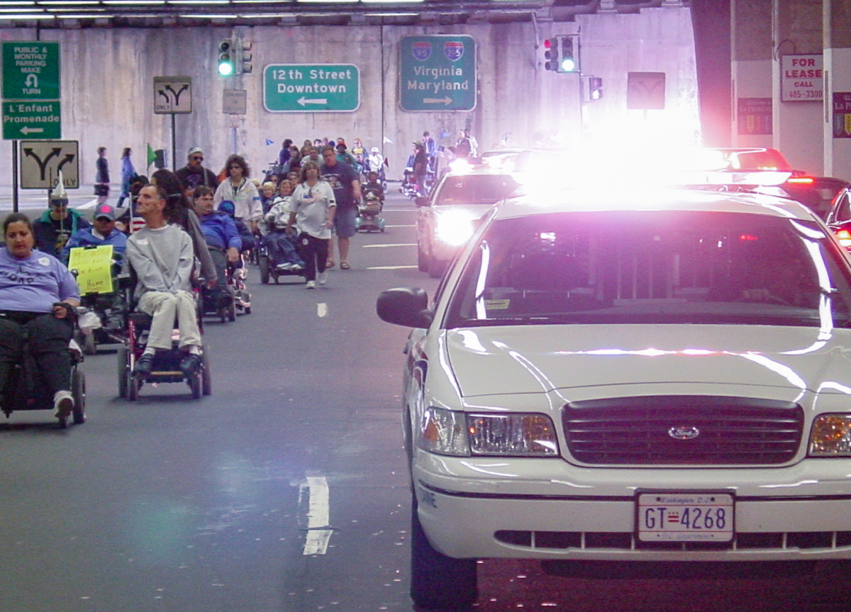 A police car with flashing lights takes a lane with people using wheelchairs in the other lane.