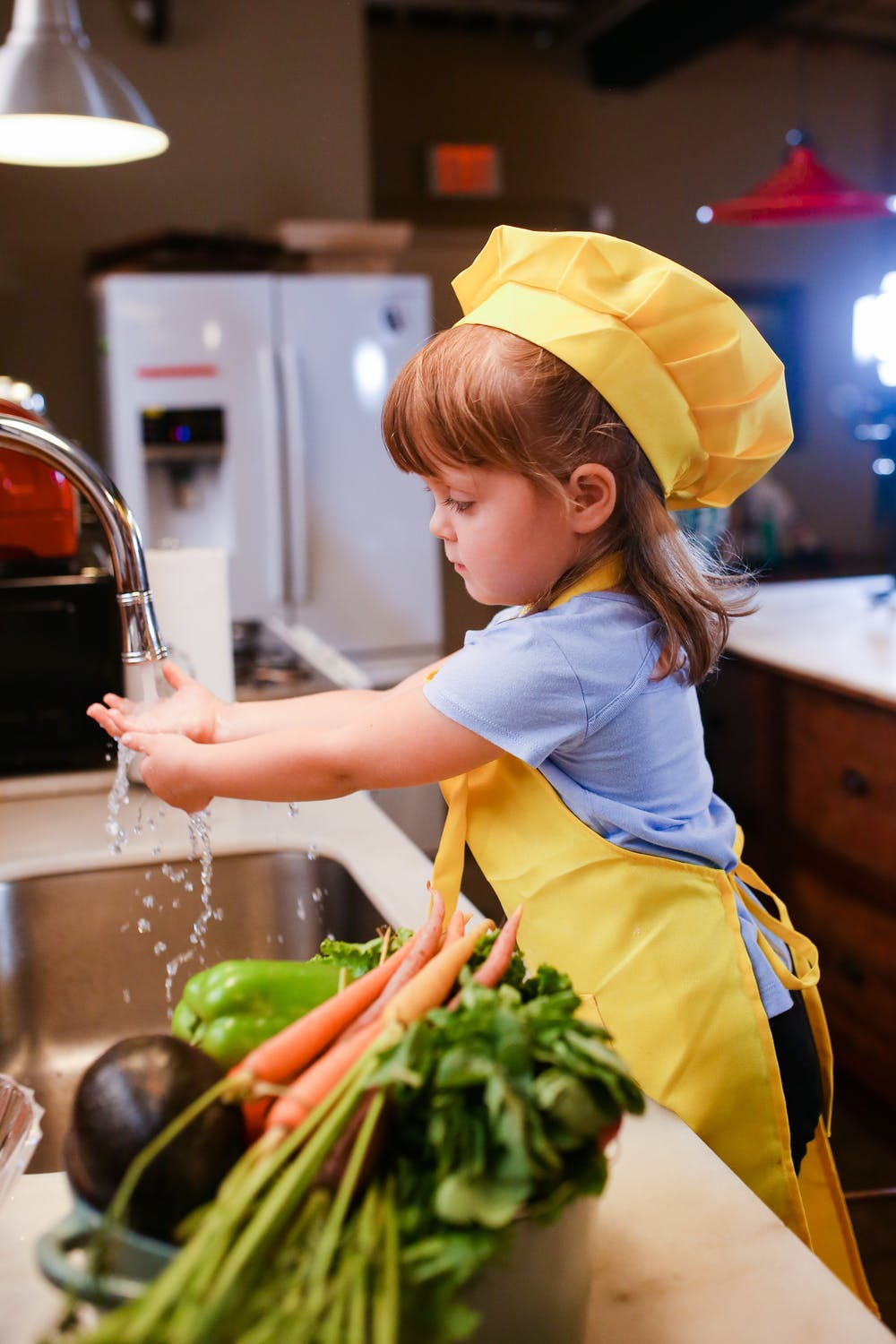 a kid wearing a yellow chef hat, light blue t-shirt, and yellow apron, while washing hands to prepare to cook. 