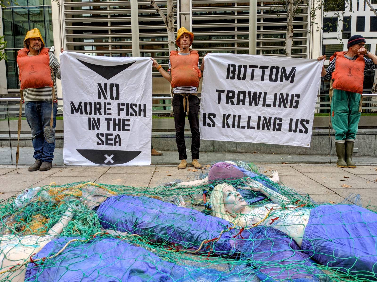 Rebels in life jackets hold sign about bottom trawling killing fish and us while below mermaids lie lifeless in netting