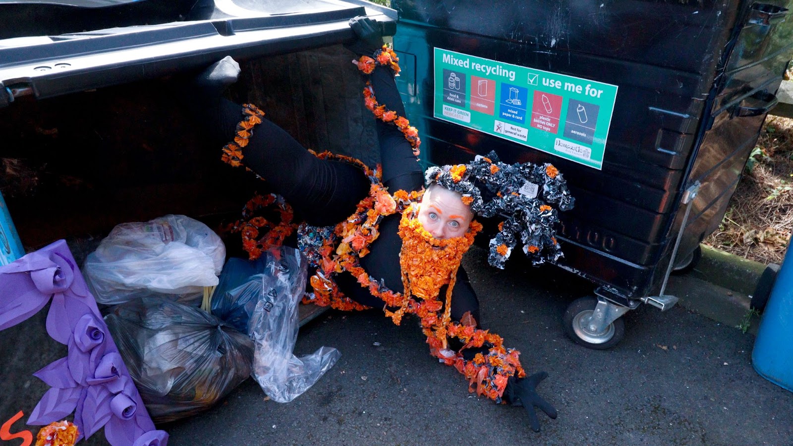 Colour photo, exterior. Kitt, a white shaven headed human, as their drag king character “Jack ‘O The Orange”. They are lying among bags of rubbish, half in, half out of a huge industrial bin, which is on its side on the street. Kitt wears a black all in one leotard covered in strings of bright orange plastic flowers. They are sporting bright orange eye brows, a Viking-esque beard made of orange plastic flowers and an antler-like headdress made of black plastic flowers. On the right hand side of the image is another industrial sized bin on which the following text is visible “Mixed recycling. Use me for” .