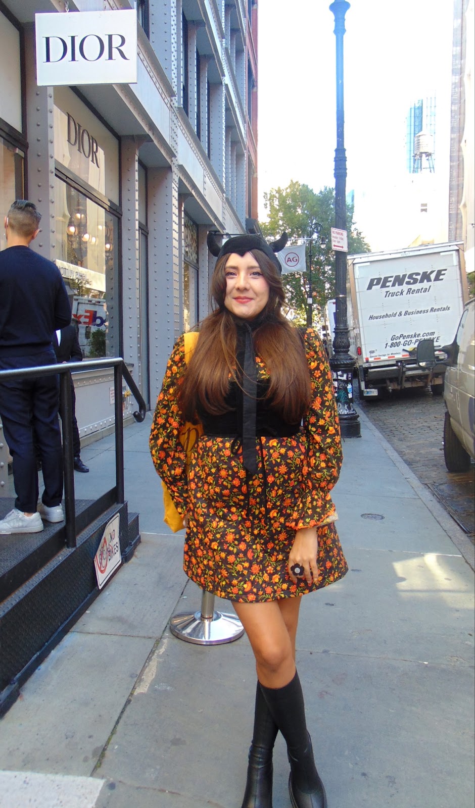 Krystal Alexis in front of the Dior store in Soho wearing a black horned hood, orange floral print dress, and knee high boots. 