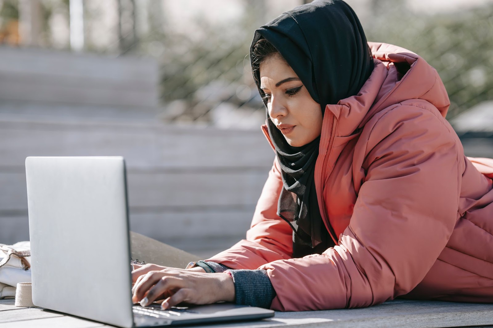 A focus shot of a young woman, wearing a hijab and a winter jacket, lying on a bench outside and working on her laptop