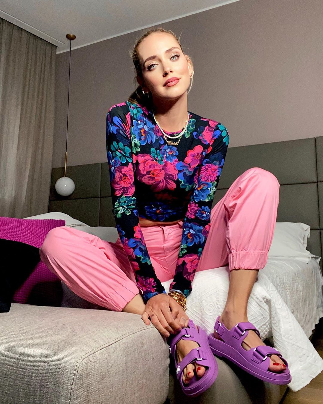 La modelo internacional Chiara Ferragni posa sentada sobre su cama mirando a cámara, mientras enseña unas sandalias de plataforma de color morado.