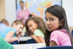 The image features a child with long, dark hair looking into the camera with a worried expression. Two other children are out of focus in the background, whispering to one another and laughing.