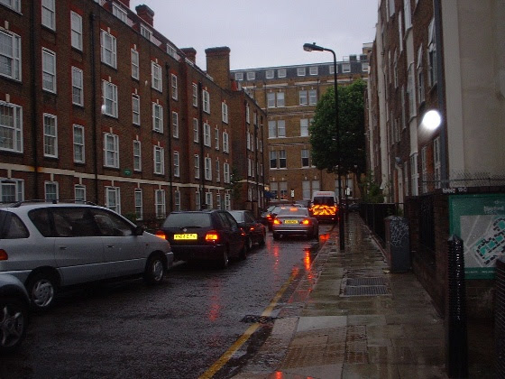 cars queuing double file on a small residential street, trying to file onto a main road in the distance on the photo