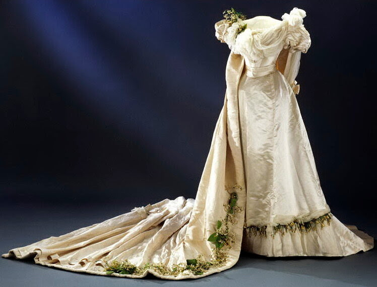 Wedding dress, 1897. Ivory atlas with white silk taffeta over bust and satin ribbon at waist. Yellow & white orange blossom and lily of the valley in wax & fabric attached to shoulder, hem, & train. Train of ivory silk rep (possibly moire) lin Wedding dress, 1897. Ivory atlas with white silk taffeta over bust and satin ribbon at waist. Yellow & white orange blossom and lily of the valley in wax & fabric attached to shoulder, hem, & train. Train of ivory silk rep (possibly moire) lin
