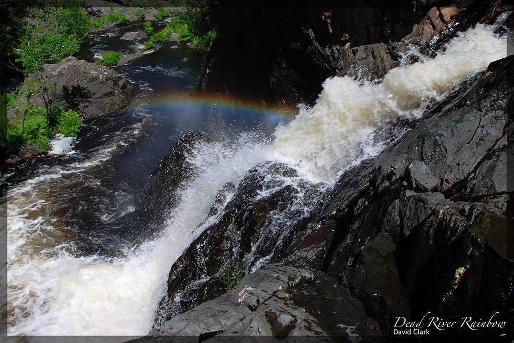 A view from the top of a rocky waterfall, looking downstream, with a rainbow visible in the mist from the fall.