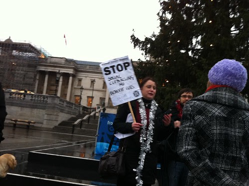 A woman standing under the tree holding up a sign saying 'stop social cleansing and benefit cuts'