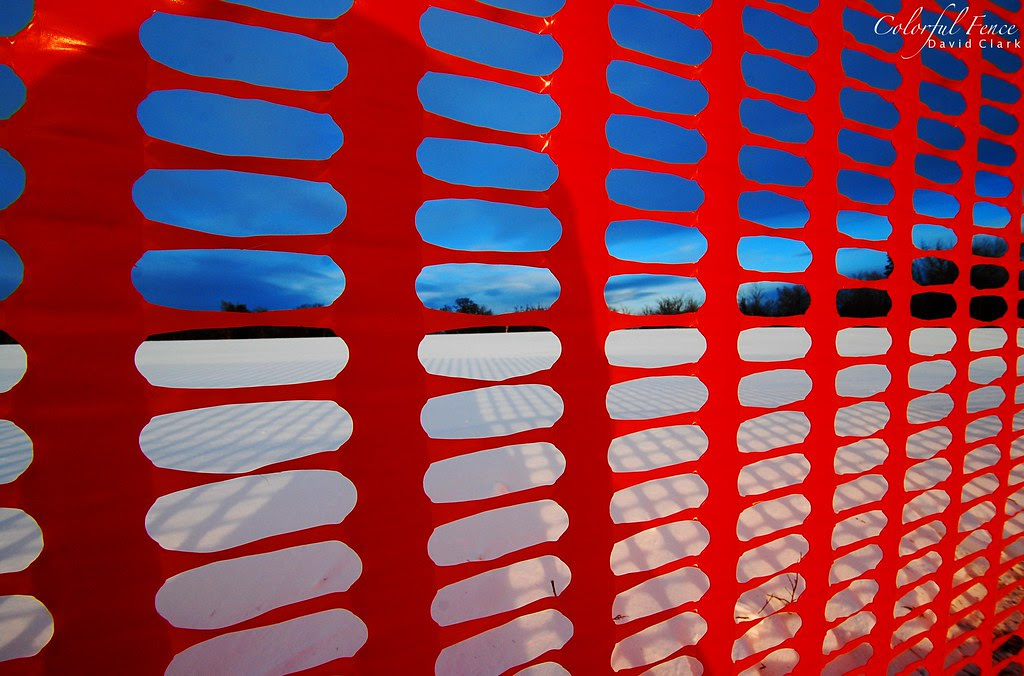 A bright orange snow fence, with shadows, snow, and blue sky behind it.