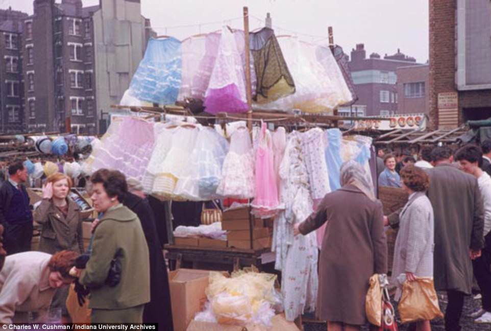 Londoners browse colourful frocks being sold at a market in Aldgate. A hot dog stand can be seen in the background to the right while a stall selling women's hats is off to the left