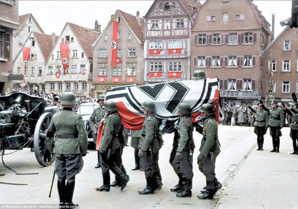 German soldiers carrying a casket of a soldier draped in a Nazi flag. People can be seen lining the streets and watching from windows of buildings adorned with Nazi propaganda. Four soldiers can be seen in the right of the picture performing the Nazi salute