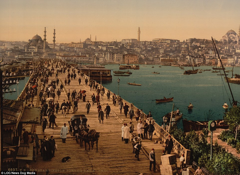 Hundreds of people walk across the Galata bridge in Constantinople, as small boats sail in the water in what was a major trade route into Europe during the Ottoman Empire