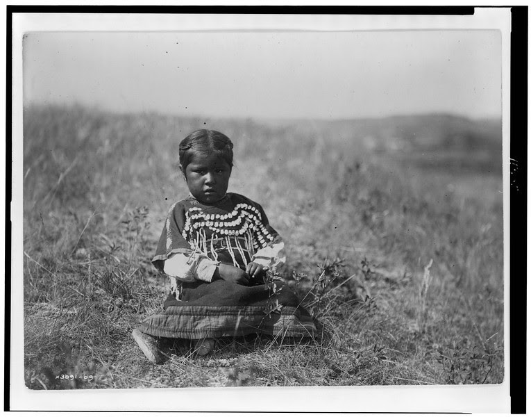 Description of  Title: Running Owl's daughter.  <br />Date Created/Published: c1910.  <br />Summary: Young Piegan girl, full-length portrait, wearing dress decorated with elk teeth, sitting on open ground, facing front.  <br />Photograph by Edward S. Curtis, Curtis (Edward S.) Collection, Library of Congress Prints and Photographs Division Washington, D.C.