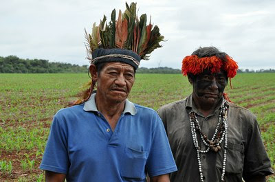 Na imagem há dois indígenas com cocares na cabeça, olhando para a câmera e em frente a uma lavoura