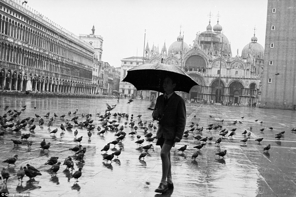 Spring showers: A schoolboy walks through the rain in Saint Mark's Square, Venice, surrounded by Saint Mark's Cathedral and the Doges' Palace, circa 1958