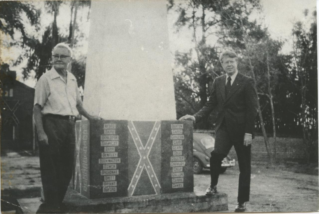 Americana resident Jim Jones stands with then-Georgia Governor Jimmy Carter alongside a commemorative monument at the Americana cemetery. Carter made the visit in 1972. Notice the Confederate insignia and names of the original ninety-four settlers of Americana etched in the based on the monument. (Photographer unknown)