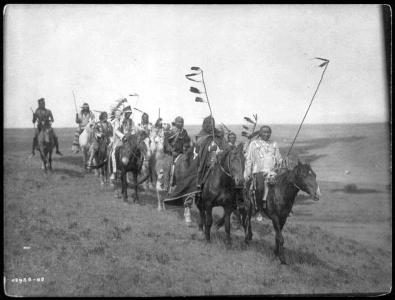 Description of  Title: On the war path--Atsina.  <br />Date Created/Published: c1908 November 19.  <br />Summary: Small band of Atsina men on horseback, some carrying staffs with feathers, one wearing a war bonnet.  <br />Photograph by Edward S. Curtis, Curtis (Edward S.) Collection, Library of Congress Prints and Photographs Division Washington, D.C.