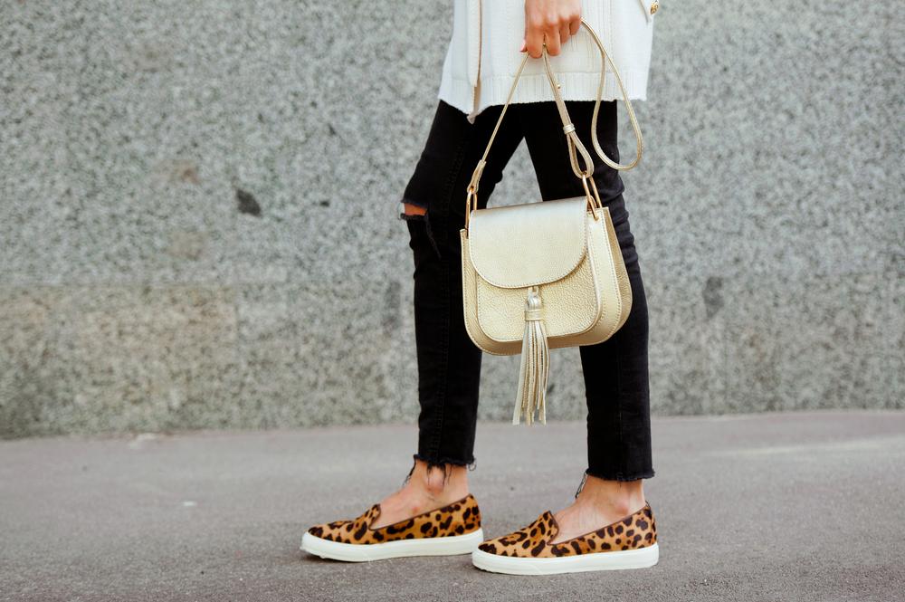 Fashionable young woman in black jeans, white cardigan and leopard shoes with gold handbag on the city streets. Fashion. Stylish .