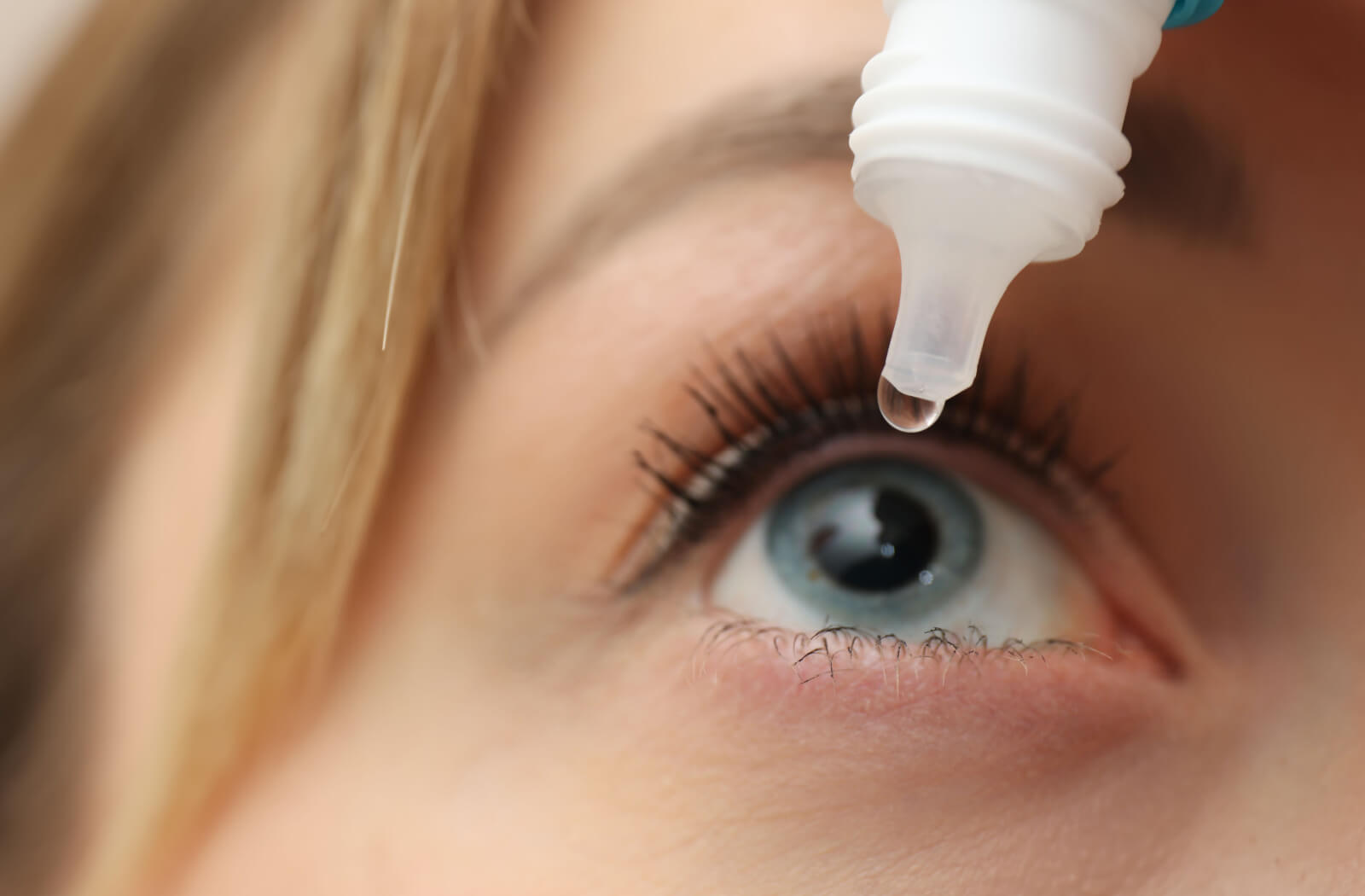 A close-up of a woman with a contact lens pouring lubricating eye drops into her right eye.