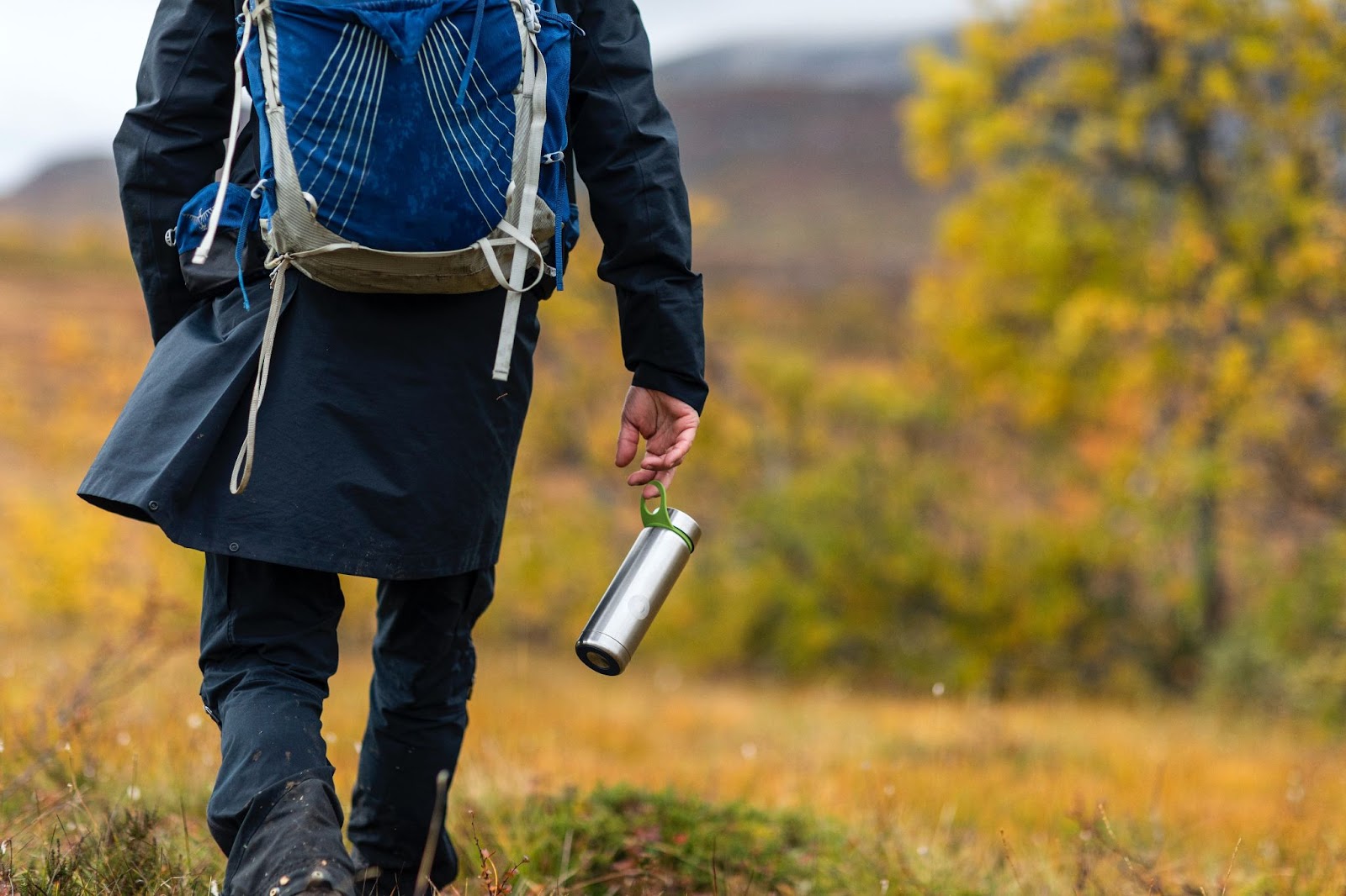 Person with a blue backpack and reusable water bottle walking over rural ground