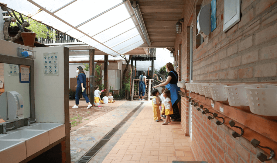 A imagem mostra um corredor, cabides para pendurar mochilas, cestos pendurados na parede. Ao fundo, crianças e adultos.