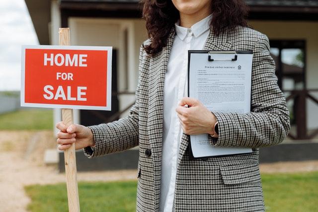 Woman in a gray jacket holding a notepad in her left hand and a home for sale sign in the right one