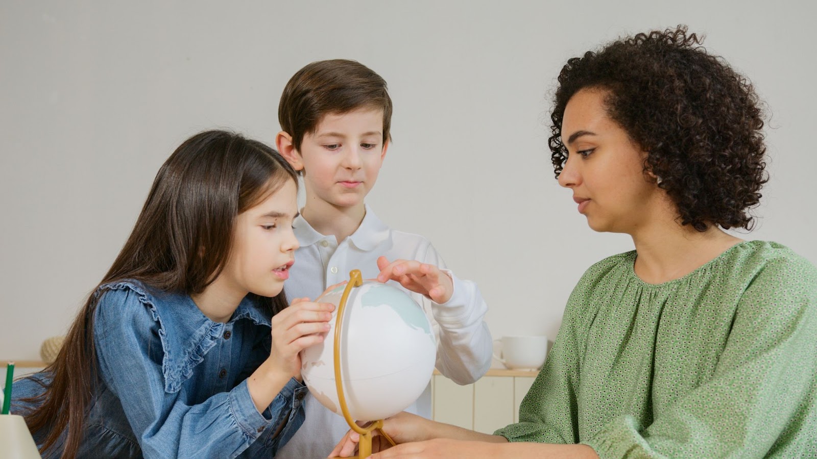 Em sala de aula, há dois alunos e uma professora segurando um globo terrestre. Da esquerda para a direita, em primeiro plano, temos uma menina branca de cabelos castanhos e lisos que usa um vestido jeans e segura na parte de cima do objeto. O menino, um pouco atrás, também é branco dos cabelos castanhos e lisos, usa uma roupa social branca e segura no globo terrestre. Ao seu lado, a professora, uma mulher negra de cabelos castanhos bem cacheados, está vestindo uma roupa verde.