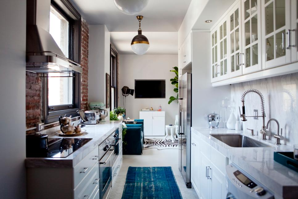 narrow galley kitchen with white lower cabinets and glass door upper cabinets. one wall framing the window has an exposed brick backsplash. a turquoise blue runner rug distracts the eye and adds a touch of style