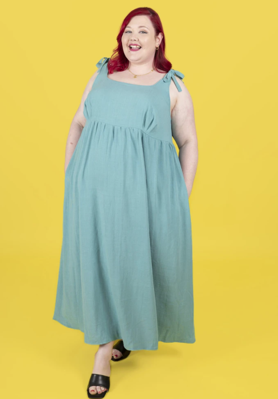 White woman with red hair wearing a seafoam green sundress, standing against a bright yellow background.