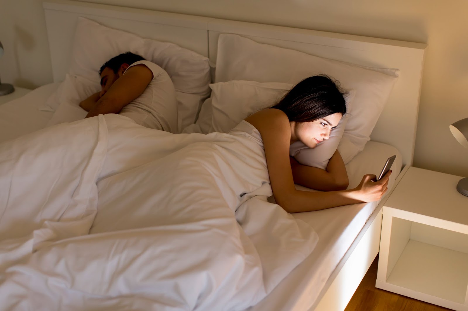 Late-night scene of a couple in bed highlighting the benefits of a motion isolation mattress, with a woman awake and using her smartphone while the man sleeps undisturbed beside her in a softly lit bedroom.