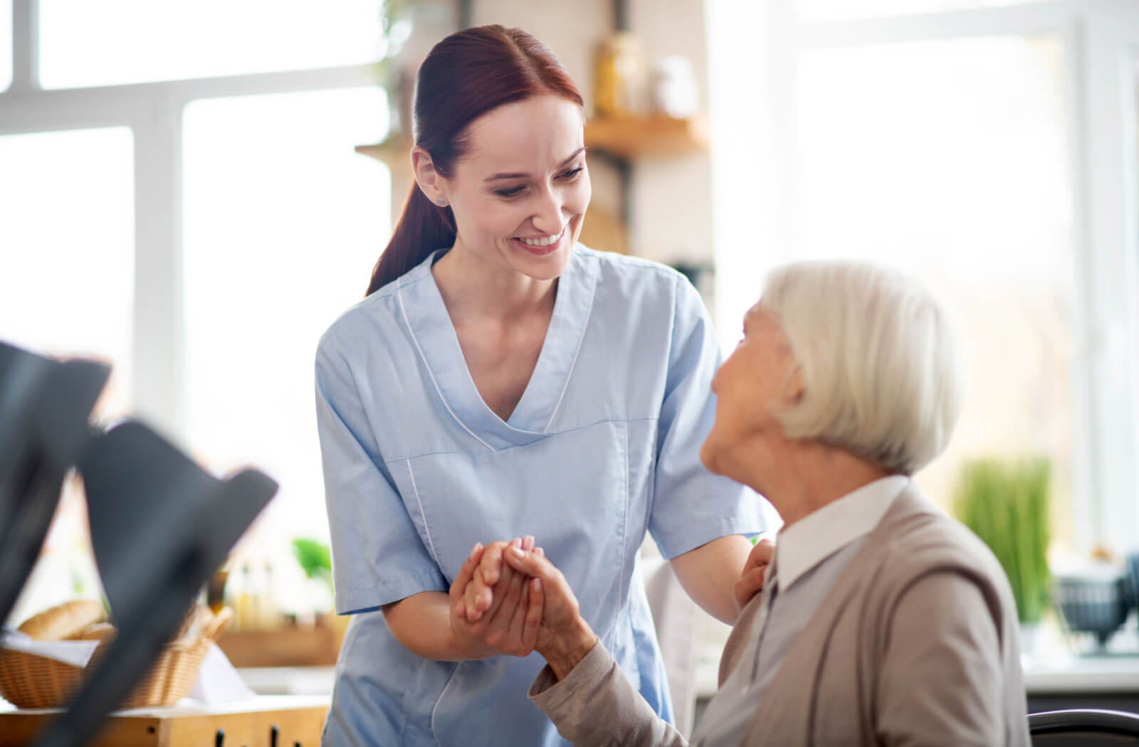 A female caregiver holding a senior woman's hand while looking at her and smiling.