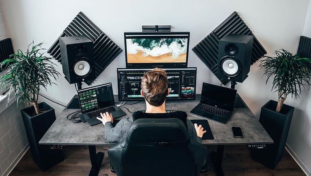 A man is working at his desk in his home office. He has dual monitors, two laptops, and high-end speakers with sound dampening panels installed on the wall directly behind them.