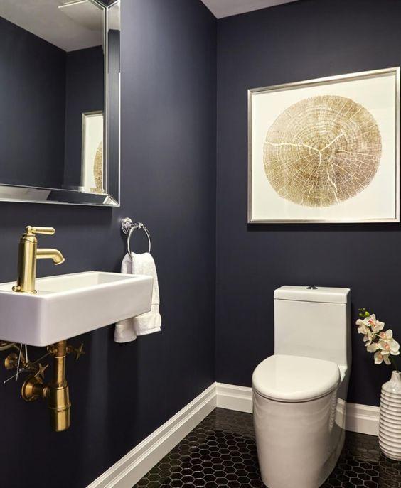 Banheiro com paredes azul escura, pia branca com torneira e encanamento dourados, espelho quadrado com moldura prata, quadro decorativo em tom neutro e piso hexagonal preto em formato pequeno