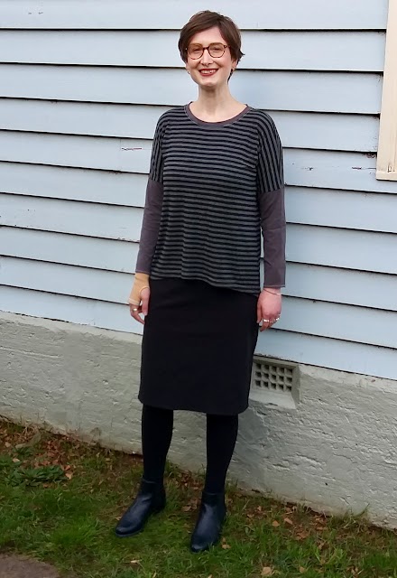 Siobhan stands in front of a weatherboard house. She wears a drop shoulder knit tee with grey/black striped body and concrete grey neckband and sleeves, with black straight knee length skirt, black leggings and ankle boots. She is smiling.