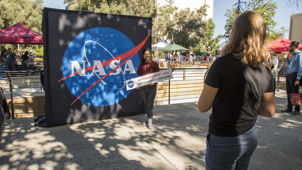 A young girl stands in front of a large NASA logo as another girl takes her photo.