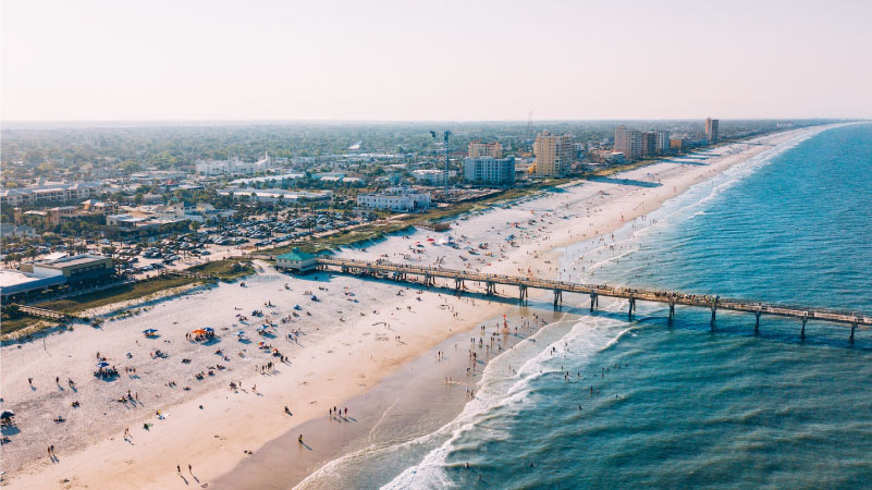 Aerial view of Jacksonville Beach and its pier. The water is a beautiful blue and people are enjoying a sunny day at the beach. 