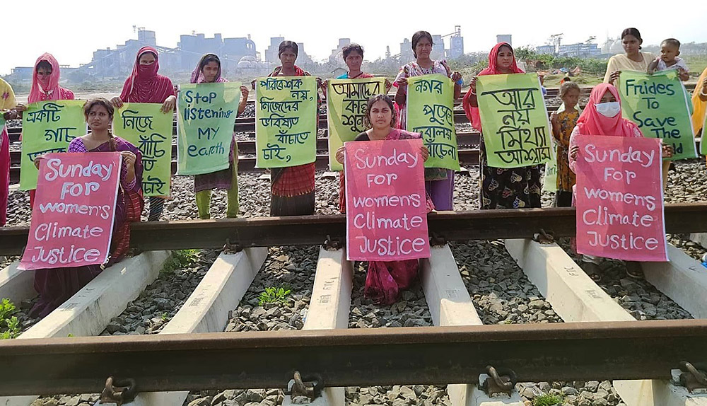 Women stand in a line along a railway track holding signs in Bengali and English about women's climate justice