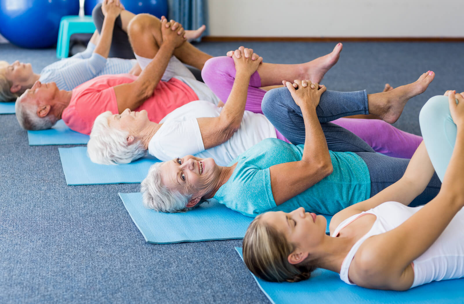 A group of seniors lay on their backs on yoga mats. They are all doing a pose where they are holding their left leg towards their chest with bent knee. One senior woman smiles at the camera.