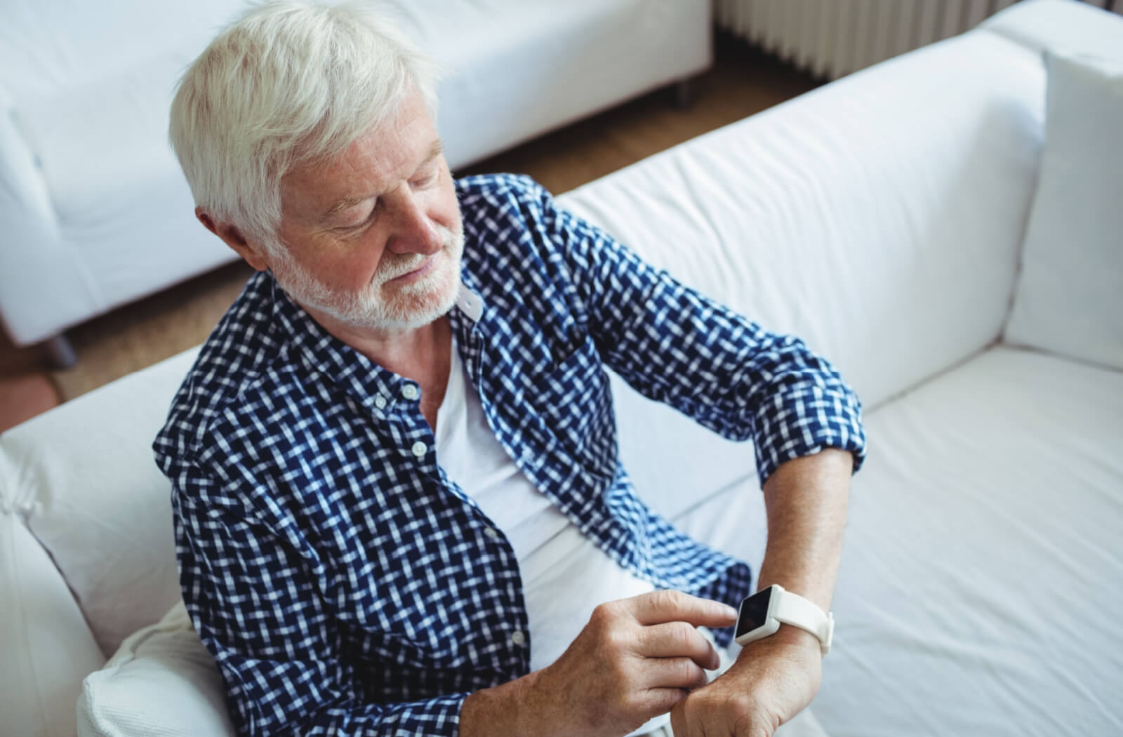 A senior man sitting on a couch and adjusting the time on his smartwatch.